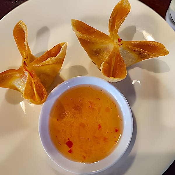 Two golden-brown crab rangoons with a side of sweet chili dipping sauce on a white plate.