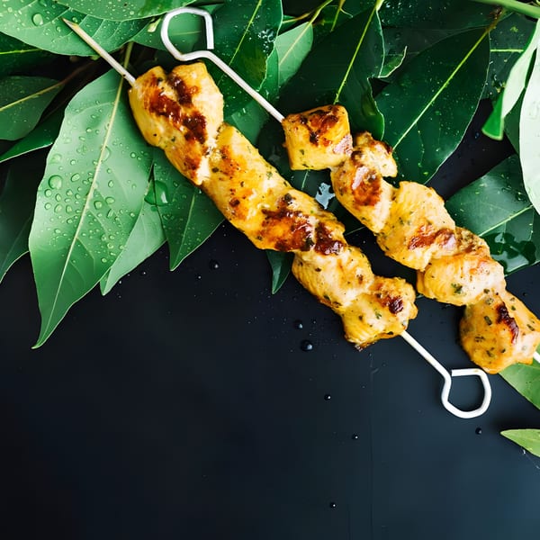 Two skewers of grilled chicken tikka on a bed of green leaves with water droplets.